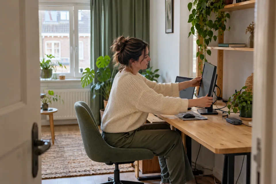 Vrouw richt een laptop en los beeldscherm in op een kleine lichte thuiswerkplek.